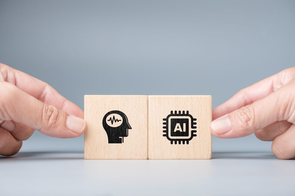 Close-up of two human hands holding wooden blocks with brain and AI chip icons symbolizing collaboration between human intelligence and artificial intelligence for innovation and creative application