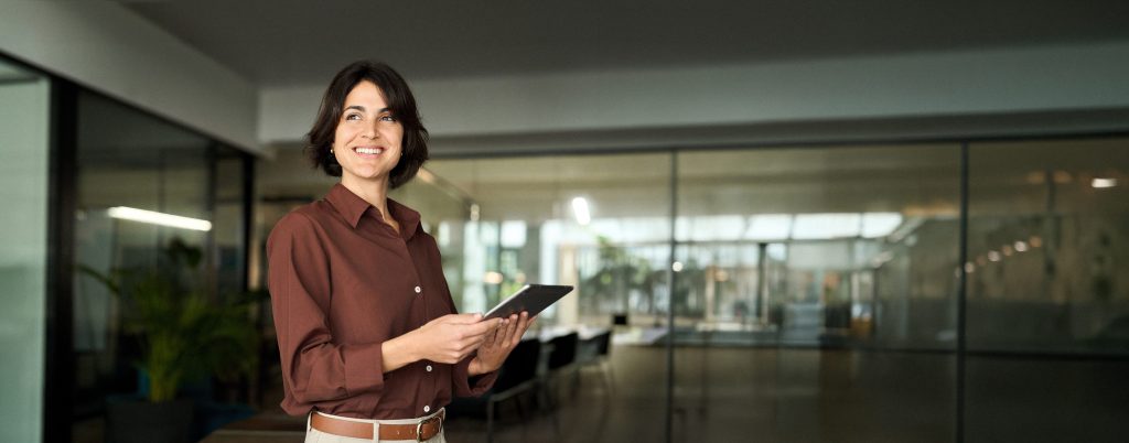 Happy young Hispanic professional business woman using tab standing in office. Happy businesswoman manager female executive leader holding tab at work looking away dreaming of future successful career